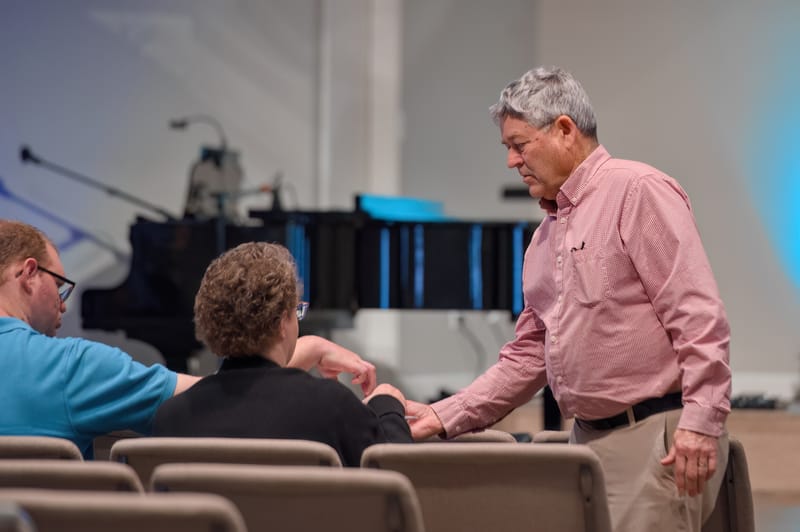An older member in a pink shirt greeting seated members before the Sunday service begins.