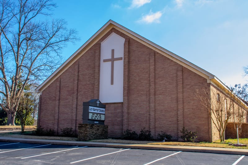 The Lucy Baptist Church building and front parking lot on a sunny day, with the church sign at the corner.