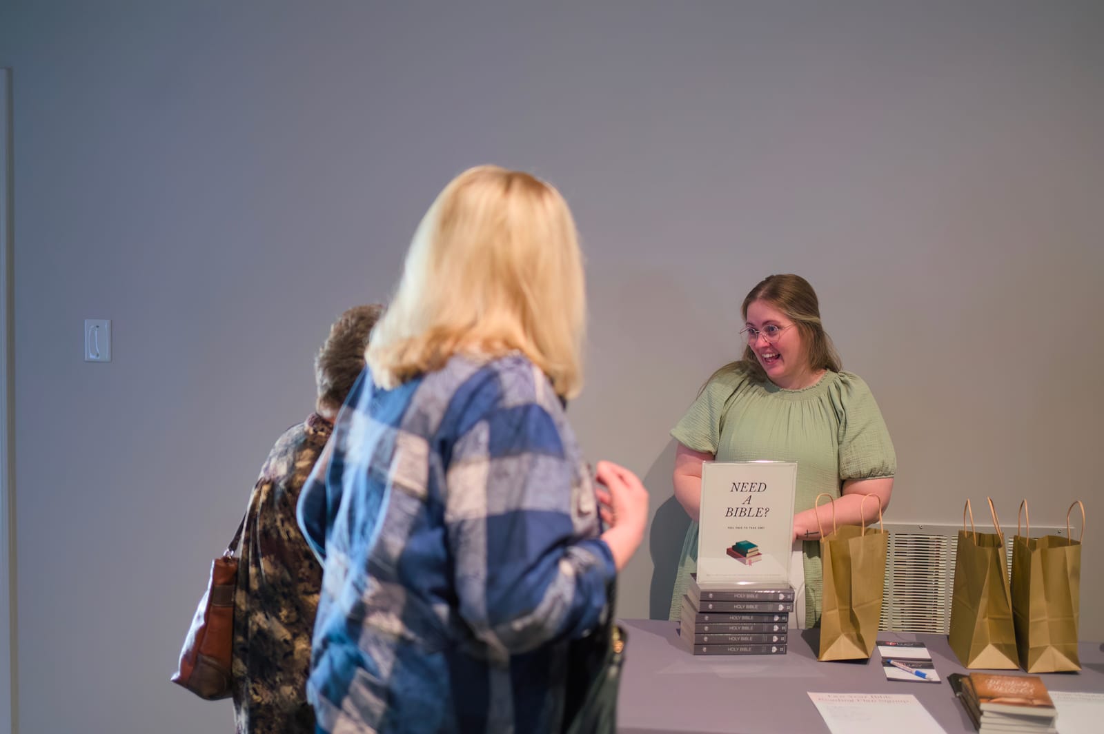 A volunteer at the welcome table offering a Bible to visitors, beside a 'Need a Bible?' sign and gift bags.